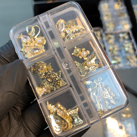 A person wearing a black glove holds a clear plastic organizer with six compartments, each filled with Dynamic Nail Supply's 30pcs (Random Shapes) nail charms in gold and silver. The intricate designs feature pearls, rhinestones, and various shapes. Additional shimmering charms can be seen in the background.