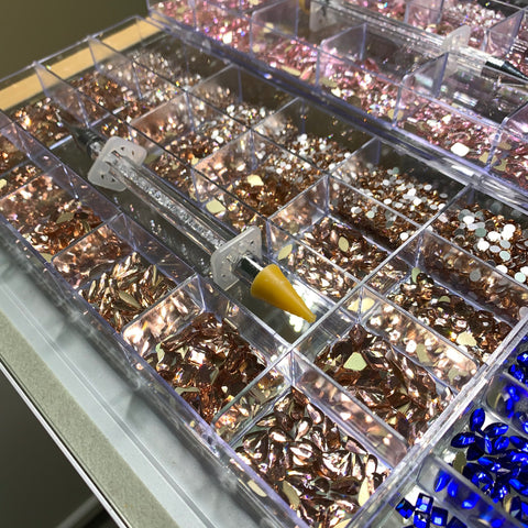 A Bling Bling set from Dynamic Nail Supply includes a segmented tray filled with shiny gemstones in vibrant hues like pink, brown, and white. A precision rhinestone picker featuring a yellow tip is conveniently nestled inside for easy selection of stones. Additionally, blue gemstones are included to add an extra sparkle to the bottom right section of the tray.