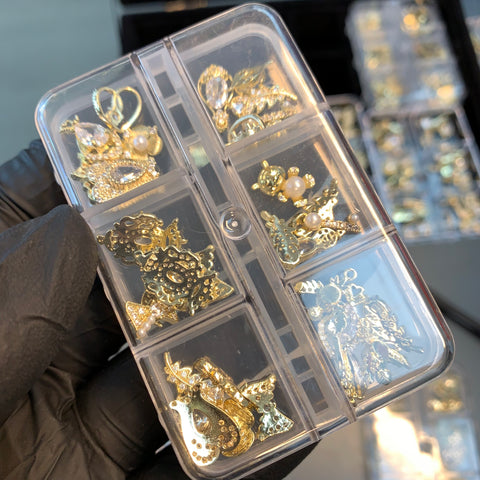 A gloved hand holds a transparent box containing assorted gold jewelry pieces, featuring charms with pearls and intricate designs. The blurred background features similar boxes, likely filled with 30pcs of randomly shaped nail charms for nail art design and decoration from Dynamic Nail Supply.