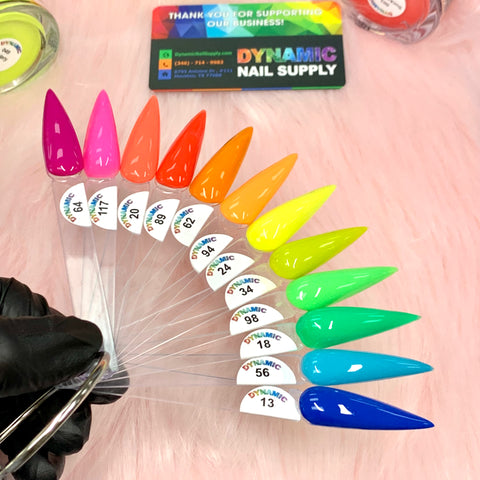 A ring of vibrant nail polish swatches from the "2024 Summer Acrylic Collection" by Dynamic Nail Supply showcases non-toxic, vivid colors like purple and pink, fanning out over a pink fluffy background. A black-gloved hand holds the ring, promising long-lasting nails. A business card and supplies complete this artistic display.