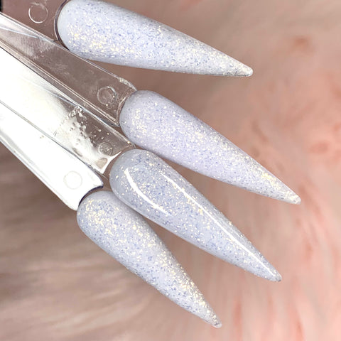 In this image, three elongated stiletto-shaped artificial nails shine with a glossy lavender finish and a hint of glitter shimmer. Created using Dynamic Nail Supply's 538 - Levitation Dream - Dynamic Acrylic Powder, these nails are elegantly arranged on a transparent holder against a softly blurred peach-toned background.