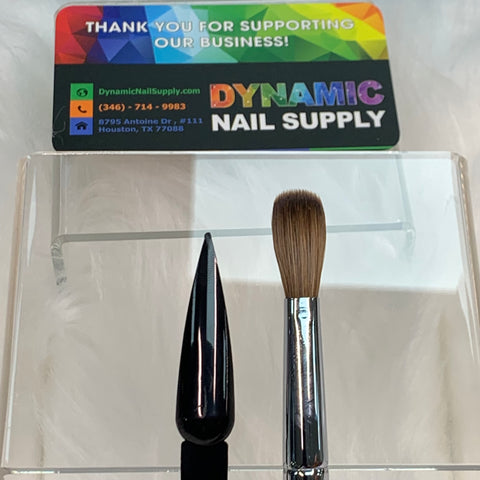 The display showcases two nail art brushes from Dynamic Nail Supply, one with a sleek black handle and the other being the Dynamic Kolinsky Hair Brush – featuring a crimped brush with a metallic silver handle and brown bristles. In the background, there's a colorful business card for Dynamic Nail Supply displaying contact details, all set against a fluffy white backdrop.