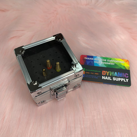 A compact, open silver Drill Bit Holder from Dynamic Nail Supply, crafted from aluminum and featuring a black foam interior, securely contains two metallic nail drill bits—one in gold and the other in bronze. Nearby is Dynamic Nail Supply's vibrant business card placed against a soft pink textured background.