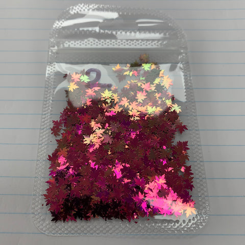 A small plastic bag from Dynamic Nail Supply, filled with their Fall leaf glitter/Maple leaves sequin for nails art design, showcases shiny and iridescent pink and purple maple leaf shapes. Resting on lined notebook paper, these color-shifting glitters catch the light in various hues, with the sealed top contrasting beautifully against the orderly lines of paper beneath.