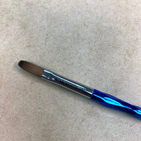 A crimped brush from Dynamic Nail Supply, featuring a blue textured handle and a metallic shaft, rests on a textured beige surface. The bristles are crafted from brown pure Kolinsky hair, with slightly rounded tips. "DYNAMIC" is engraved on the metallic part of this Dynamic Kolinsky Brush, making it ideal for acrylic nail application.