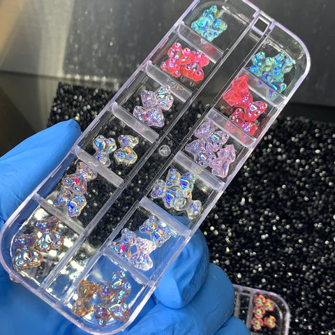 A hand wearing blue gloves holds a clear plastic organizer from Dynamic Nail Supply, featuring small compartments filled with colorful, iridescent decorative nail gems in shapes like stars, moons, gummy bears, and butterflies. The background displays a textured black surface adorned with additional scattered gems.