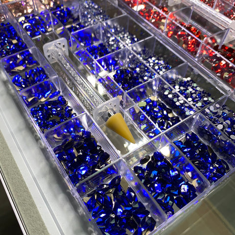 A dazzling collection of sparkling gemstones from Dynamic Nail Supply is neatly arranged in a transparent organizer. The Bling Bling set, featuring 3000 pieces each of vibrant rose, red, and blue gems in assorted shapes and sizes, surrounds a rhinestone picker tool at the center. The light dances off these vibrant hues, creating an eye-catching display from above.