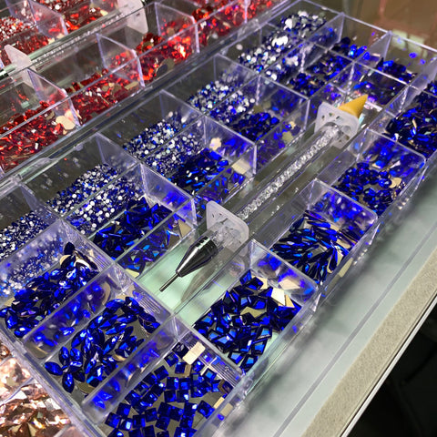 A display case from Dynamic Nail Supply, featuring the Bling Bling set, showcases compartments filled with vibrant rhinestones in shades of rose, red, blue, and gold. The set's rhinestone picker tool elegantly rests diagonally across the compartments, its clear handle and pointed tip perfectly highlighting the stones' shimmer.