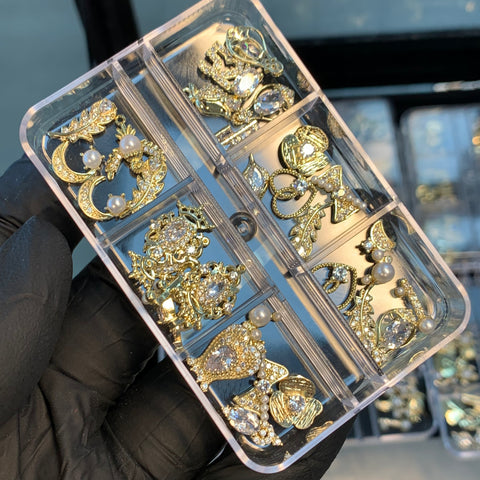 A gloved hand holds a transparent organizer filled with six compartments containing 30pcs of Dynamic Nail Supply's decorative gold and silver nail charms. Each compartment features ornate designs with pearls and sparkling rhinestones, showcasing intricate patterns in random shapes perfect for nail art or jewelry making.