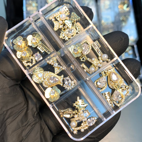 A gloved hand holds a clear plastic container featuring six compartments, each brimming with an assortment from Dynamic Nail Supply's 30pcs Random Shapes Nail Charms. These exquisite metal charms feature intricate designs embellished with pearls and rhinestones, resembling shapes such as hearts, crowns, and animals.