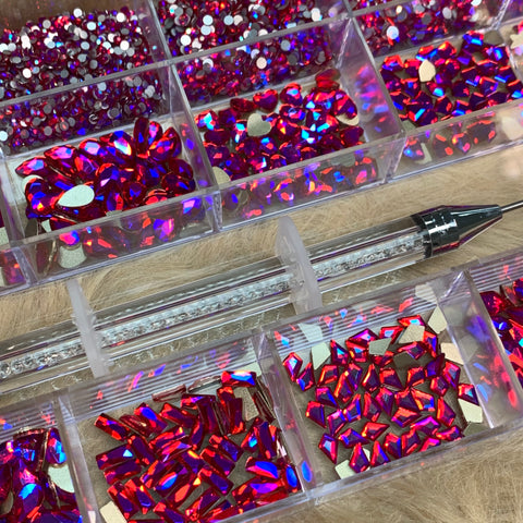 Introducing the Dynamic Nail Supply [iridescent magenta] Rhinestones Set—a stunning collection featuring vibrant red and pink gemstones in 14 unique big shapes and 6 round shapes, all neatly organized in a 20-cell clear compartment box. This assortment is ideal for any nail supply enthusiast. Accompanying the set is an elegant iridescent magenta rhinestone picker tool with a textured grip, gracefully placed across the boxes on a light brown surface.