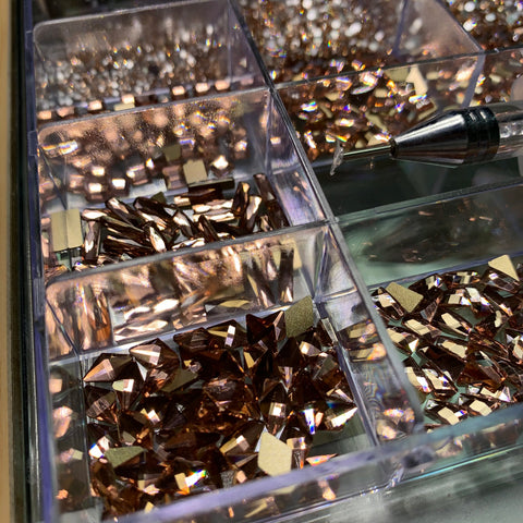 A close-up of the Bling Bling set from Dynamic Nail Supply showcases various metallic brown and rose-gold crystals neatly arranged in a divided container. Positioned near the upper right section is a rhinestone picker with a pointed tip, ready to organize the shimmering gems. Each compartment contains sparkling crystals that reflect light, creating a dazzling effect.