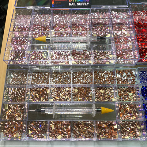 A vibrant display of nail art supplies from Dynamic Nail Supply's Bling Bling set showcases assorted gems in shades of rose, red, blue, and gold, stored in neatly organized compartments separated by type and size. Two empty syringes with yellow tips, ideal for use as rhinestone pickers, rest atop the arrangement.