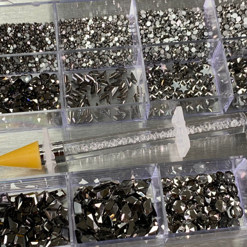 A crystal-clear container from Dynamic Nail Supply showcases a stunning collection of gun metal rhinestones, featuring both black and silver hues, in a variety of sizes and 14 big shapes alongside 6 round shapes. A yellow-handled picker with a tapered tip lies elegantly against the edge of the case. The metallic backdrop accentuates the charm of these gleaming stones.