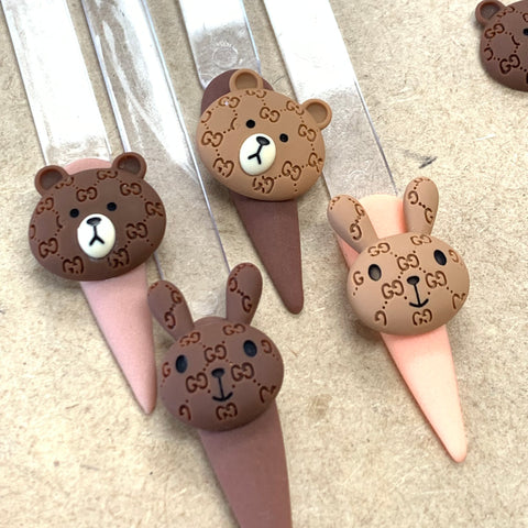 Close-up of four delightful nail art charms, featuring adorable animal designs on a beige background. Two charms display brown bear faces with small black eyes and noses, while the other two showcase rabbits with long ears. Perfect for your Instagram-worthy collection, each face includes decorative patterns like tiny bows or flowers. These are part of the Logo pattern Brown & Cony charms for nails art design by Dynamic Nail Supply.