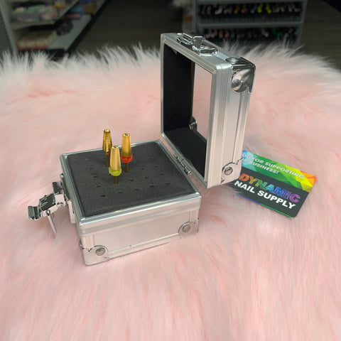 A small Drill Bit Holder from Dynamic Nail Supply, crafted in silver aluminum with a padded black interior, elegantly displays four gold and red nail drill bits in an upright position. The open container box features a shiny exterior and a hinged lid, all set on a soft pink, fluffy surface. A colorful business card accompanies the scene.
