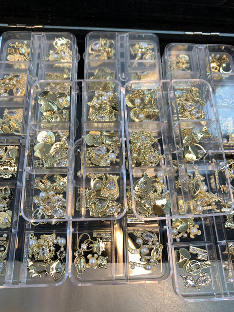 A collection of jewelry pieces in gold tones, including earrings and pendants with random shapes like stars and teardrops, some embellished with pearls and rhinestones, is neatly organized in clear plastic storage boxes. These pieces are arranged in individual compartments alongside Dynamic Nail Supply's 30pcs (Random Shapes) Nail Charms for nail art design and decoration.