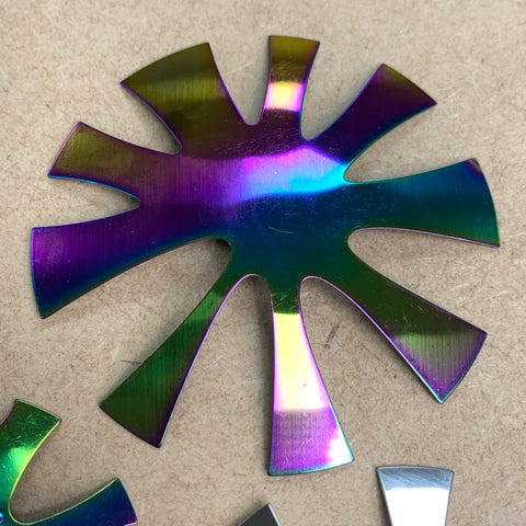 A metallic, iridescent French Nails Cutter by Dynamic Nail Supply, designed to resemble a French Smile Line Cut with uneven, outward-reaching arms, rests on a plain beige surface. Its acrylic trimmer-like finish reflects a spectrum of colors such as purple, blue, green, and yellow.