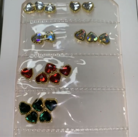 The Pre-made Nail Charms from Dynamic Nail Supply feature a clear plastic sheet filled with heart-shaped gemstones, ideal for nail designers. The top row includes five silver gems, while the second and third rows contain five and three red gems respectively. The bottom row consists of six green gems. Each gemstone is elegantly outlined with a gold border—perfect for creating stunning nail designs.