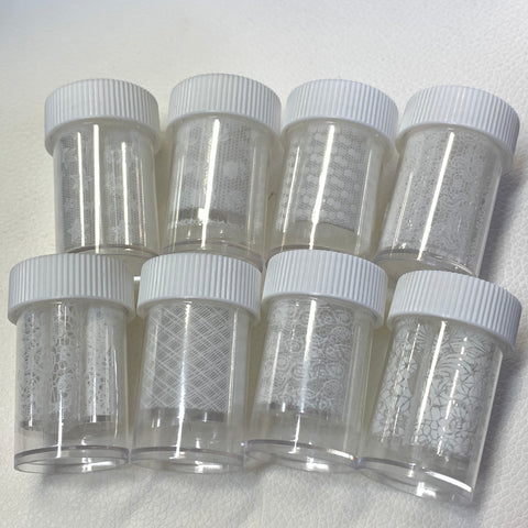 On a white textured surface, seven small clear plastic jars are displayed, each containing designer foils with intricate white patterns suitable for nail art. These jars from Dynamic Nail Supply feature style #001 and offer a total of 8 rolls, each 2 feet long. The arrangement is in two rows with four jars on top and three on the bottom, all topped with white screw-on lids.