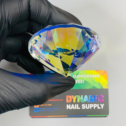 A black-gloved hand holds a large, 6cm K9 crystal diamond from Dynamic Nail Supply, known for its multifaceted brilliance ideal for display and decoration. Behind the diamond, a colorful business card with a vibrant rainbow design and small contact details completes the scene. This stunning display piece beautifully reflects shades of blue and yellow.