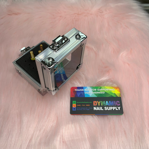 A sleek Drill Bit Holder, crafted from aluminum with a glass lid, displays an assortment of gold and black drill bits elegantly resting on a fluffy pink surface. Next to it, a vibrant business card for Dynamic Nail Supply, adorned with rainbow accents and contact information, adds a splash of color.