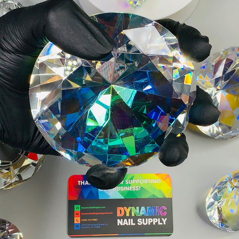 A gloved hand holds a large, faceted 10cm (4") Display Diamond made of K9 Crystal, beautifully reflecting rainbow colors. Below, a vibrant business card reads Dynamic Nail Supply with contact details and a message of appreciation. Other similar gems are elegantly scattered across the surface, serving as table decorations.