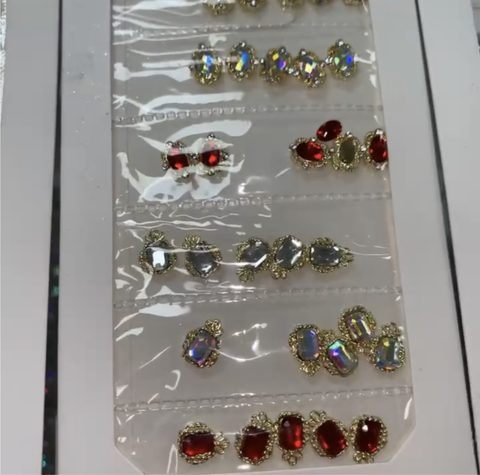 A collection of the Pre-made Nail Charms / Gems for Nails Designer by Dynamic Nail Supply is elegantly displayed in a plastic package. It features exquisite gold-framed, multi-faceted red and iridescent charms, each meticulously arranged in neat rows and separated by plastic seams, set against a crisp white background.