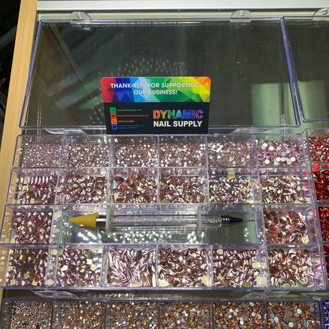 A transparent display case showcases compartments filled with pink and clear nail art rhinestones in various shapes. The set comes with a rhinestone picker pen that has a yellow tip for improved precision. A vibrant sign from Dynamic Nail Supply expresses gratitude for supporting their brand at dynamicnailsupply.