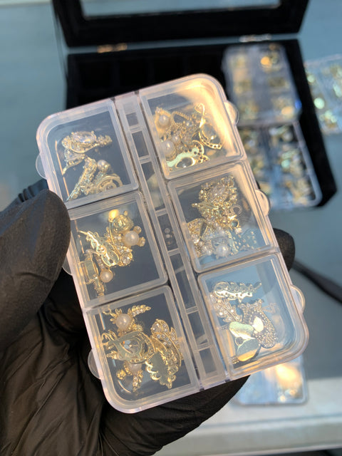 A gloved hand holds a transparent plastic container labeled "30pcs (Random Shapes) Nail Charms for Nails art design and decoration" by Dynamic Nail Supply. The container features six compartments, each filled with various small, intricate metallic nail charms that glint like gold and include designs such as hearts, stars, and random shapes. A tray of similar containers is visible in the background.
