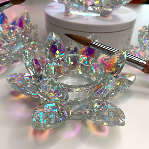 A CRYSTAL LOTUS DAPPEN DISH from Dynamic Nail Supply, shaped like a delicate flower, reflects vibrant rainbow colors when crafted from iridescent rainbow crystal glass. Nearby, there is a round white container and a brush with a wooden handle that add an artistic touch to the scene. The bright light enhances the colorful display by illuminating the dazzling facets of the crystal dish.