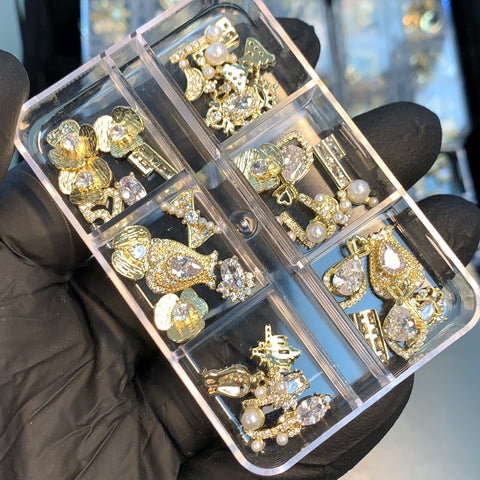 A gloved hand holds a transparent box featuring 30 pieces of gold-colored nail charms from Dynamic Nail Supply, ideal for nail art design and decoration. The assortment includes bows, hearts, and random shapes, each adorned with shiny rhinestones and small pearls. Different styles are neatly organized in each compartment of the box.