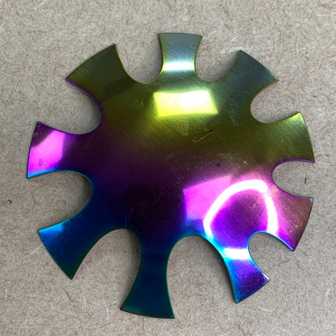 A French Nails Cutter from Dynamic Nail Supply, designed with a metallic, iridescent gear shape and eight rounded, fan-like protrusions, reminiscent of the Easy French Smile Cut V Line tool, rests on a beige surface. It reflects shades of purple, blue, green, and yellow, creating a circular shimmer on its lower right section.