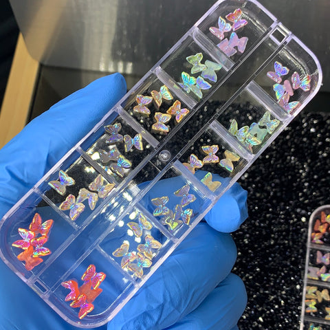 A gloved hand holds a clear plastic container featuring multiple compartments, each filled with iridescent nail art pieces shaped like Dynamic Nail Supply's Gummy Butterflies. The butterflies shimmer in an array of colors, including pink, purple, and blue—reminiscent of tiny gummy butterflies against a dark background.