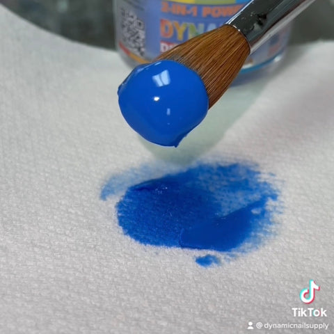 A brush dipped in Dynamic Acrylic Dipping Powder S 007 - Solid Blue (Cobalt) hovers above a white textured paper towel, where a small drop of the product has spread, suggesting intricate nail art possibilities. In the background, a container from Dynamic Nail Supply is also visible, with the TikTok username @dynamicnailsupply displayed in the corner.