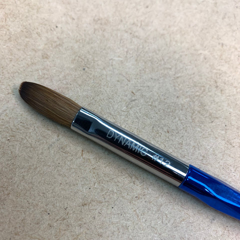 A close-up of the [Crimped] Dynamic Kolinsky Brush, featuring a blue handle and metallic silver ferrule, showcases its pointed tip bristles ideal for acrylic applications. The ferrule is inscribed with "DYNAMIC #12," set against a light brown, textured background that resembles wood or cork.