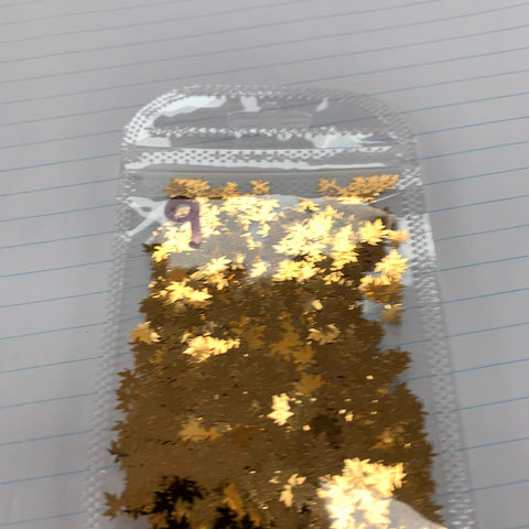 A small clear plastic bag containing Dynamic Nail Supply's Fall Leaf Glitter – Maple Leaves Sequin for nails art design – sits atop a lined notebook page. Reminiscent of fall leaves, the color-shifting glitters are densely packed towards the bottom of the bag, ideal for nail art designs. The top is sealed with a white pattern along the edge.