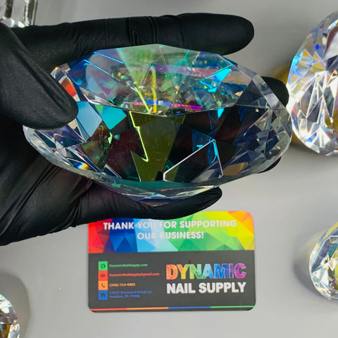 A hand in a black glove holds a large, multicolored K9 crystal with rainbow reflections. Below is a business card from Dynamic Nail Supply featuring a colorful geometric design and contact information. Similar 10cm (4") Display Diamonds from the brand, shaped for decoration, are partially visible on the sides, making them perfect for table displays.