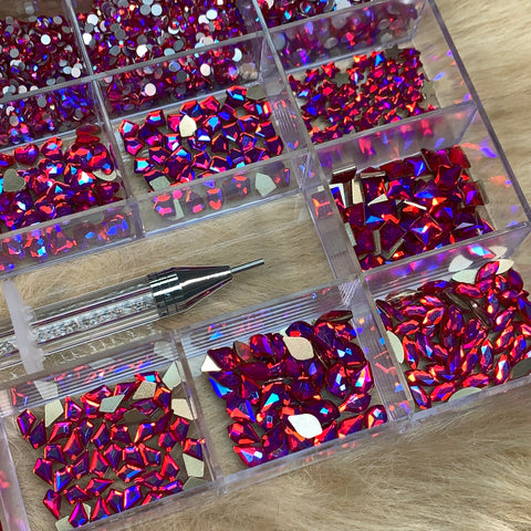 An iridescent magenta rhinestones set from Dynamic Nail Supply is beautifully arranged inside a transparent 20-cell organizer, showcasing gemstones in 14 big shapes and 6 round shapes. Alongside, a nail art tool with a pointed end and rhinestone-filled handle is gracefully positioned across one section. The setting rests on a soft beige surface, ideal for any nail supply enthusiast.
