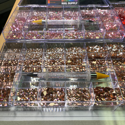 An impressive collection of rose gold nail art embellishments is beautifully organized in transparent compartments. Two rhinestone picker pens featuring yellow tips are positioned diagonally across the arrangement. A partially visible sign reads "Dynamic Nail Supply," suggesting their captivating Bling Bling set collection, which includes 3,000 pieces each of rose, red, blue, and gold embellishments.