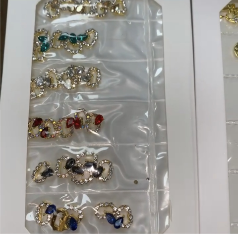 A collection of Pre-made Nail Charms / Gems for Nails Designer by Dynamic Nail Supply is showcased, with various colored stones arranged in rows on a white background. Each piece features intricate designs resembling gold earrings, with stones in shades of blue, red, green, and clear. The charms are encased in clear plastic packaging to highlight their ornate details.