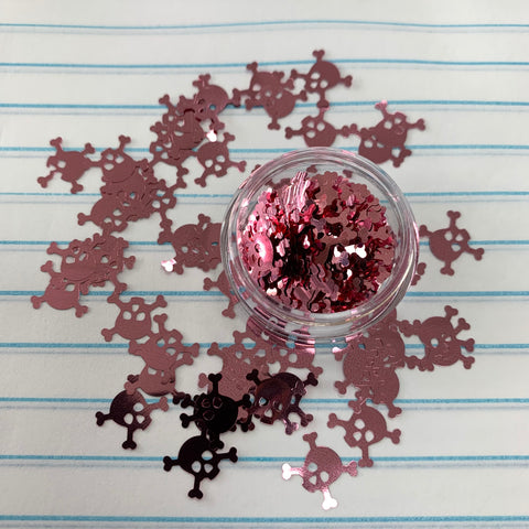 A small container from Dynamic Nail Supply, filled with clear metallic pink glitter in the shape of skulls and crossbones, is tipped over on a background of blue-lined paper. Ideal for Halloween nails, the scattered confetti creates an eerie charm reminiscent of acrylic powders on a tiny canvas.