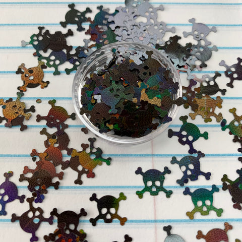 A small container of Skull Shape Glitter from Dynamic Nail Supply features metallic, multicolored turtle-shaped confetti positioned on lined notebook paper. The playful mix includes a quirky twist with skull-shaped glitter in green, blue, red, and gold hues, creating a vibrant contrast against the white and blue lines.