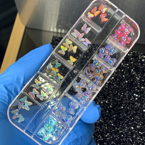 A gloved hand holds a clear plastic organizer from Dynamic Nail Supply, showcasing their colorful Gummy Butterflies and vibrant Flower Charms. The embellishments, in shades of pink, purple, and blue, are neatly arranged in separate compartments against a dark, textured background.