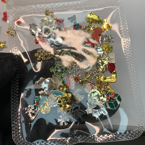 A hand wearing a black glove from Dynamic Nail Supply holds a clear plastic bag on the left side. Inside the bag, you'll find 50 Christmas charms perfect for winter nail art design, including metallic snowflakes, Christmas trees, Santa hats, reindeer, and gift boxes in gold, silver, red, and green.