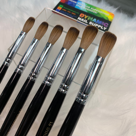 A set of seven Dynamic Kolinsky Hair Brushes, featuring black handles and silver ferrules, is elegantly arranged on a clear acrylic stand. Each brush boasts a brown bristle tip known for its flexibility in shaping acrylic. In the background, a colorful business card with "Dynamic Nail Supply" sits on a fluffy white surface.