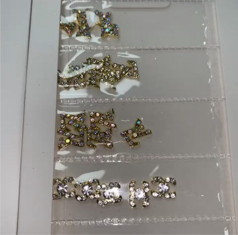 Close-up of a transparent case featuring multiple small compartments, each filled with "Pre-made Nail Charms / Gems for Nails Designer" by Dynamic Nail Supply. The gold charms exhibit a variety of designs, including bow shapes and intricate patterns embellished with sparkling rhinestones, perfect for any nail designer aiming to elevate their art on a pale surface.