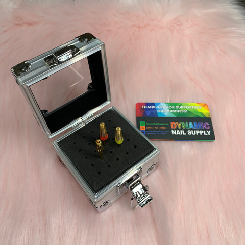 A small metallic Drill Bit Holder from Dynamic Nail Supply, crafted from aluminum with a mirror lid, is open on a soft pink surface. Inside, a foam insert holds three gold and multicolor nail drill bits securely. Nearby is a colorful business card from Dynamic Nail Supply featuring contact information and a thank you message.