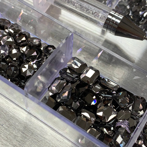 Close-up of a clear box containing the shimmering Gun Metal color Rhinestones set from Dynamic Nail Supply, featuring 14 big shapes and 6 round shapes elegantly displayed. The reflective silver surface enhances their brilliance. At the top, a diagonal placement of a clear pen with a metallic tip adds an element of intrigue to the composition.