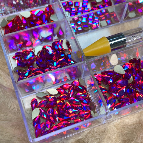 The [iridescent magenta] Rhinestones set by Dynamic Nail Supply, housed in a 20-cell box, features vibrant red and iridescent magenta rhinestones in various shapes like teardrop and square. Accompanying the set is a rhinestone picker with a yellow tip that rests atop the container. The backdrop of soft beige fabric complements any nail supply project perfectly.